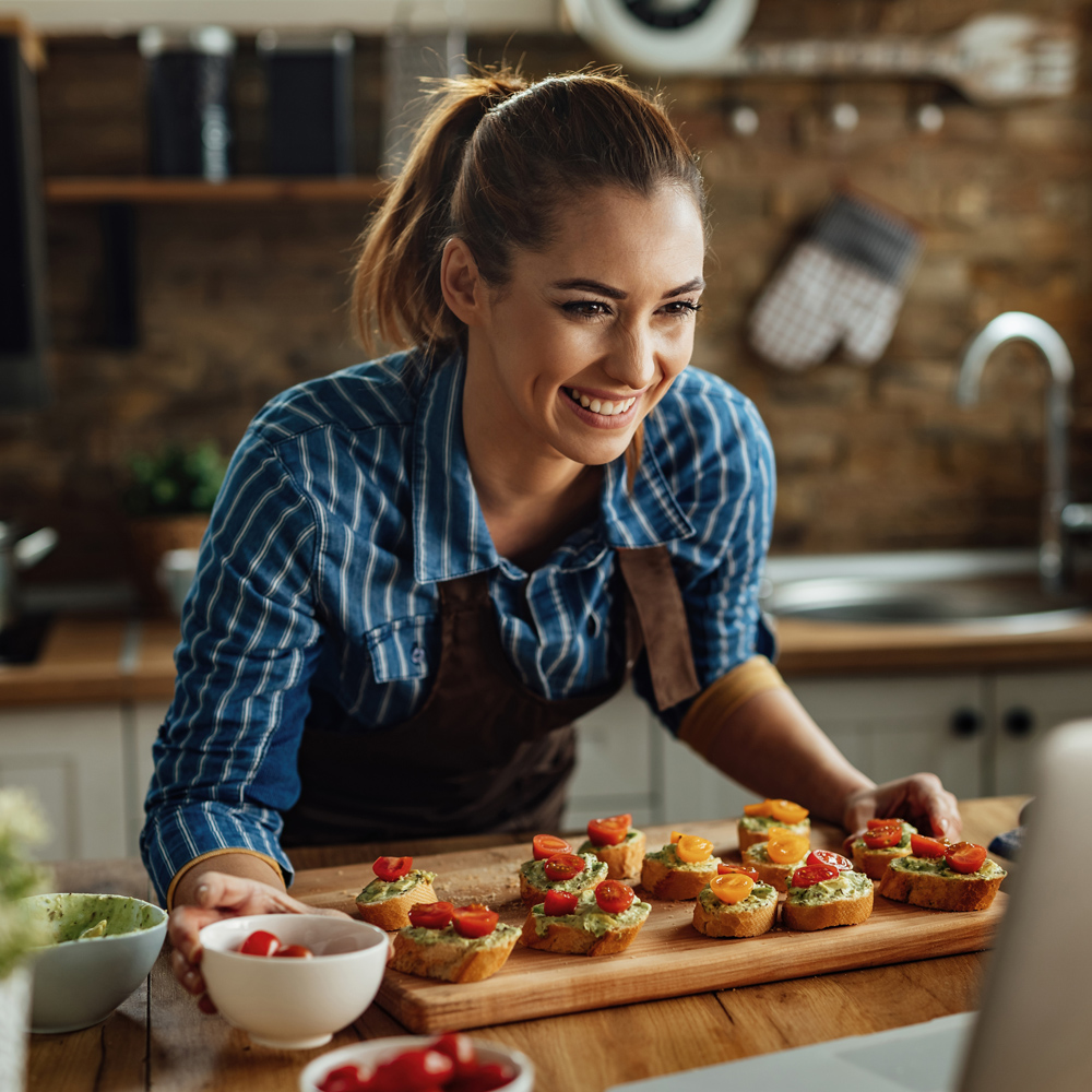 Young happy woman vlogging over laptop while preparing bruschetta with avocado and cherry tomato in the kitchen.